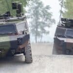 Two military armored vehicles drive on a gravel road surrounded by trees.