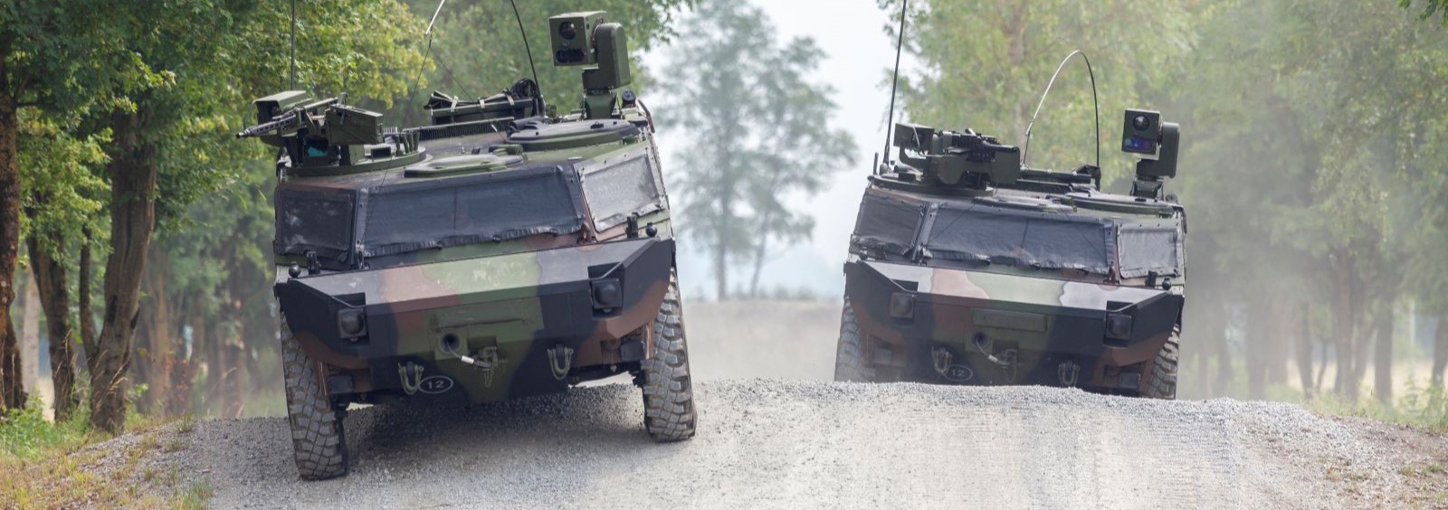 Two military armored vehicles drive on a gravel road surrounded by trees.