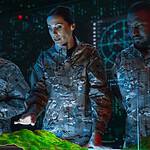 Three uniformed individuals analyze a 3D topographic map in a high-tech room with digital displays and data visuals.